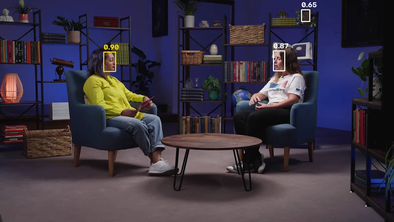 A wide frame of the interview with three boxes: a yellow one on the interviewer's face labelled 0.90, a white one on Hannah's face labelled 0.87, and a small white one on a dark shape on the top shelf labelled 0.65.